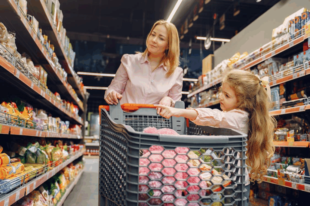 Mother and daughter shopping in a grocery store aisle with a full cart of household items.